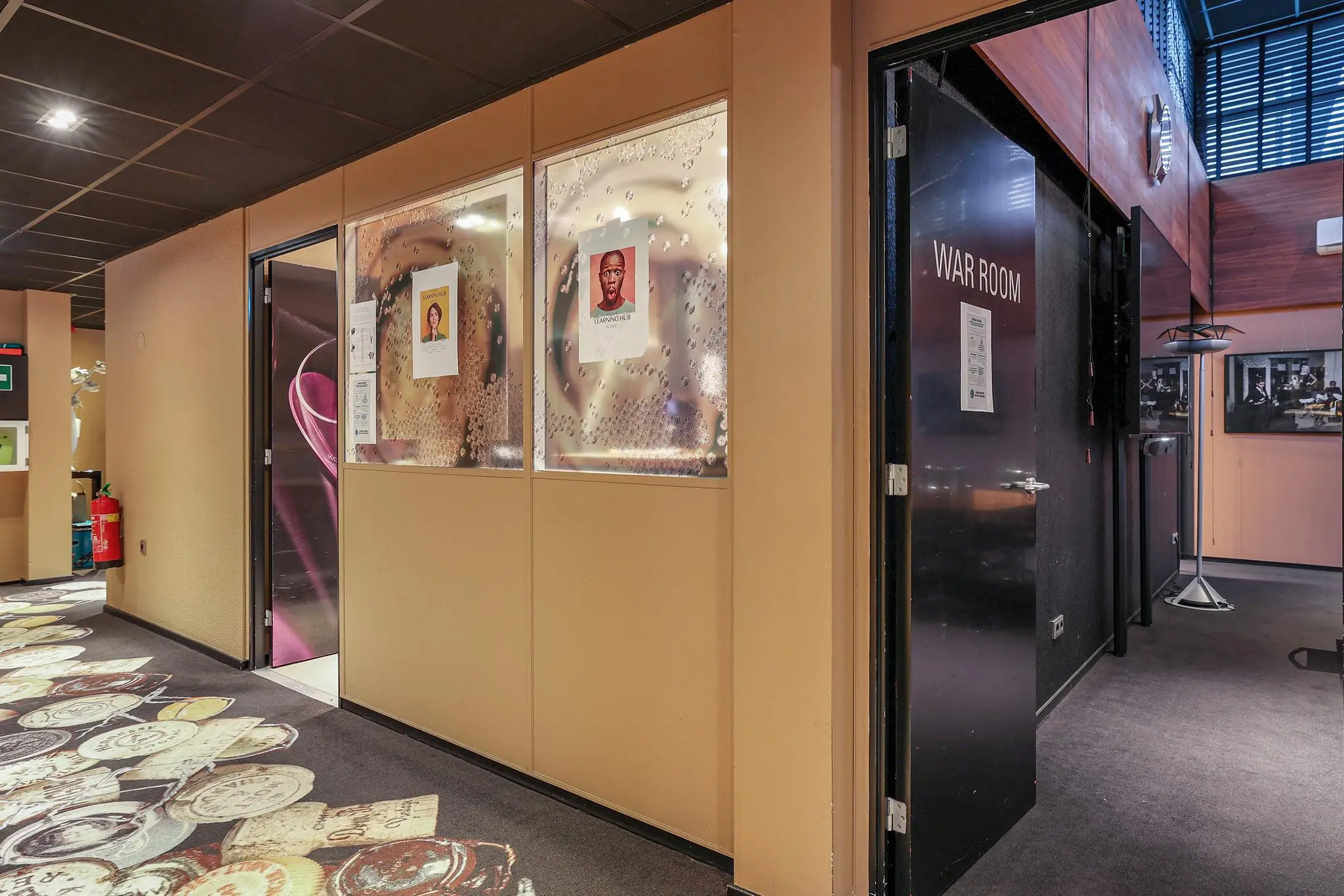 Gang in een modern kantoor met een openstaande deur naar de "War Room" en glazen wanden met posters van medewerkers.